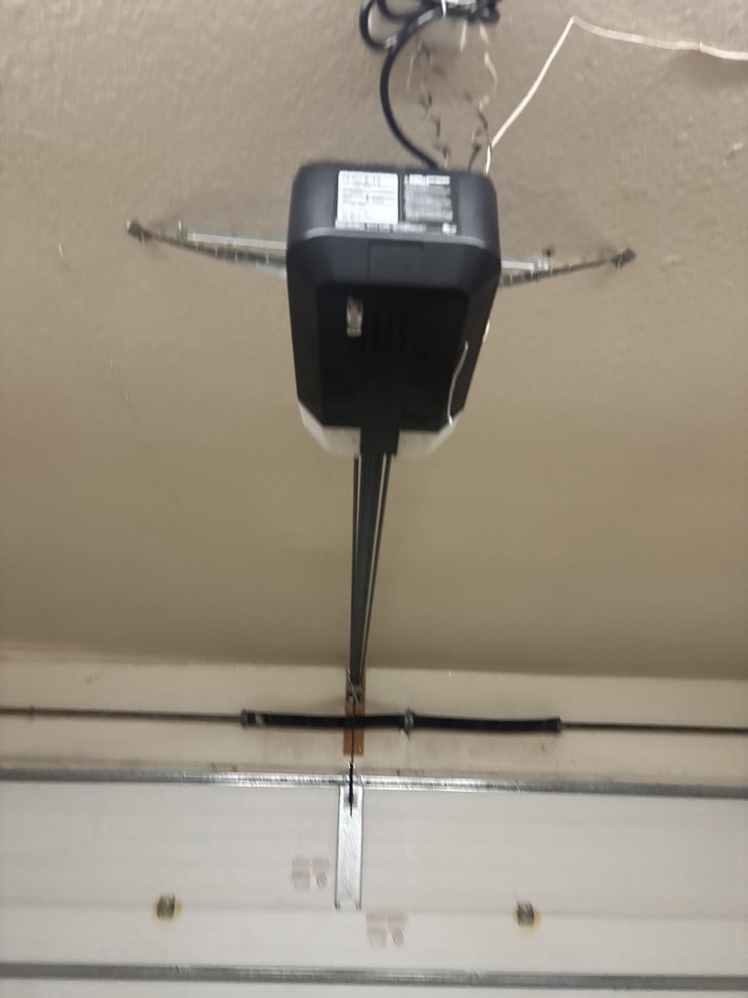 Black garage door opener motor mounted to garage ceiling with connecting wires and hardware.